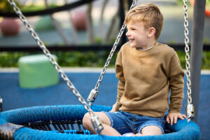 Toddler wearing Rabbit Skins Fleece Crew Neck Pullover Sweatshirt (3317) in coyote brown while playing on a swing at the playground.