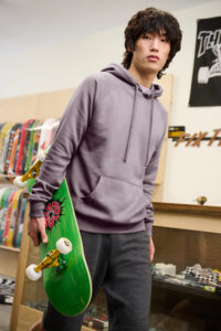 Young man wearing District Cloud Fleece Hoodie in soft lavender while holding skateboard inside skate shop.