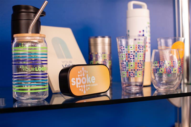 A display of Spoke Custom drinkware and hard goods—including tumblers, bottles, glassware, and a branded speaker—showcasing full-wrap sublimation and UV-printed designs.