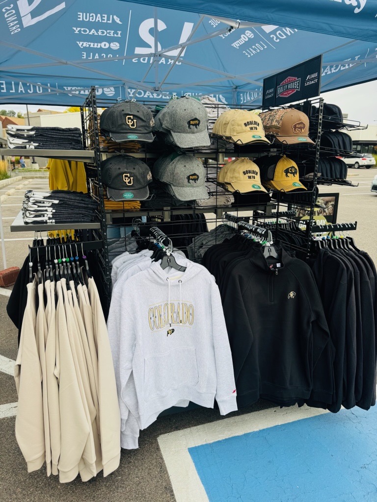 Display of Colorado Buffaloes collegiate merchandise at a Legacy Headwear game-day booth, featuring hats, hoodies, and apparel arranged under a branded tent during a campus activation event.