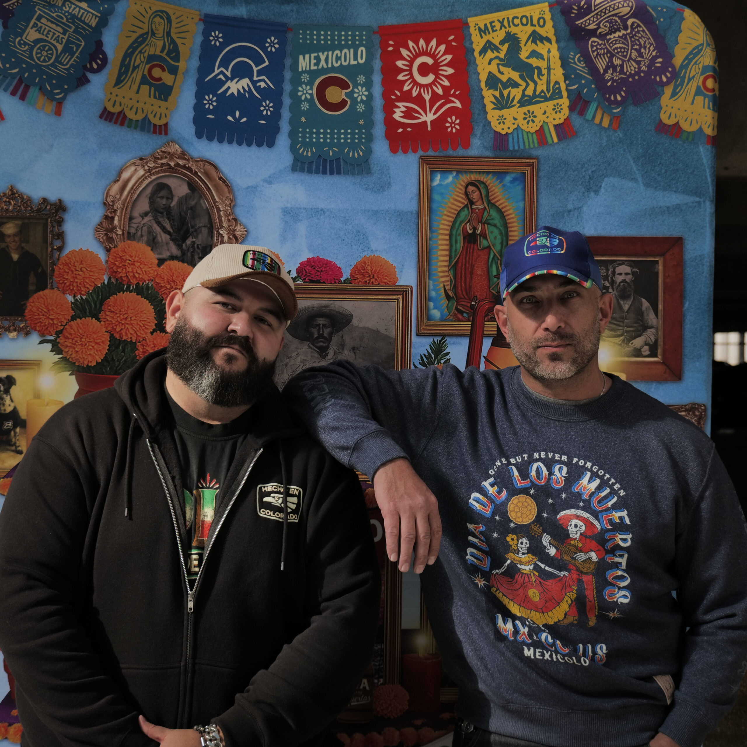 Two men wearing Mexicolo apparel stand in front of a colorful Día de los Muertos display featuring papel picado, marigolds, and framed portraits.