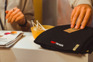 Black Atlantis Headwear Moover beanie with TEDx Venezia embroidery displayed at event table, highlighting sustainable promotional headwear partnership.