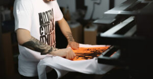 Decorator applies a direct-to-film (DTF) transfer to a custom T-shirt using a heat press for print-on-demand apparel production.