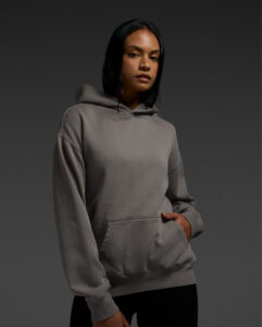 Model wearing AS Colour Women's Relax Faded Hood in faded gray, showcasing premium heavyweight fleece, relaxed fit and modern women's blank apparel styling.